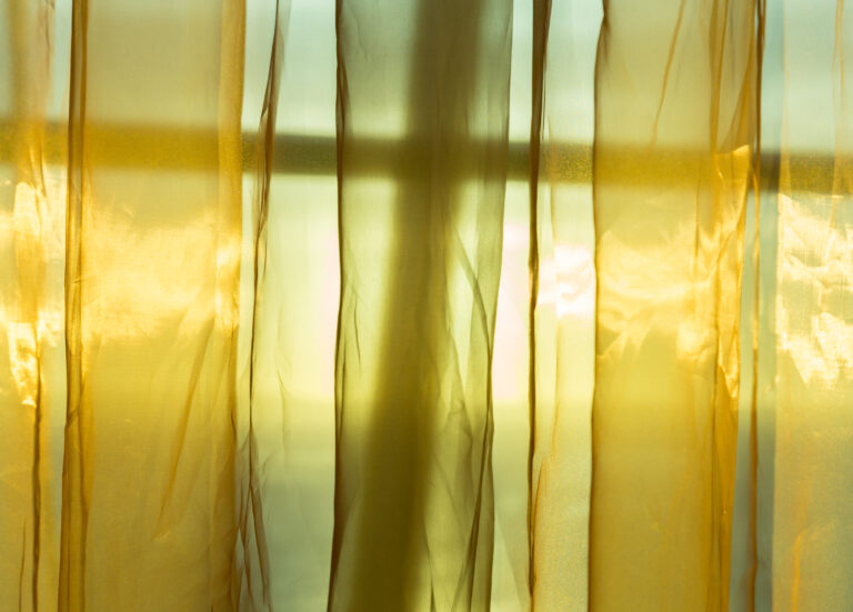 Sunlight from outside window streams into a room through thin golden silky net curtains